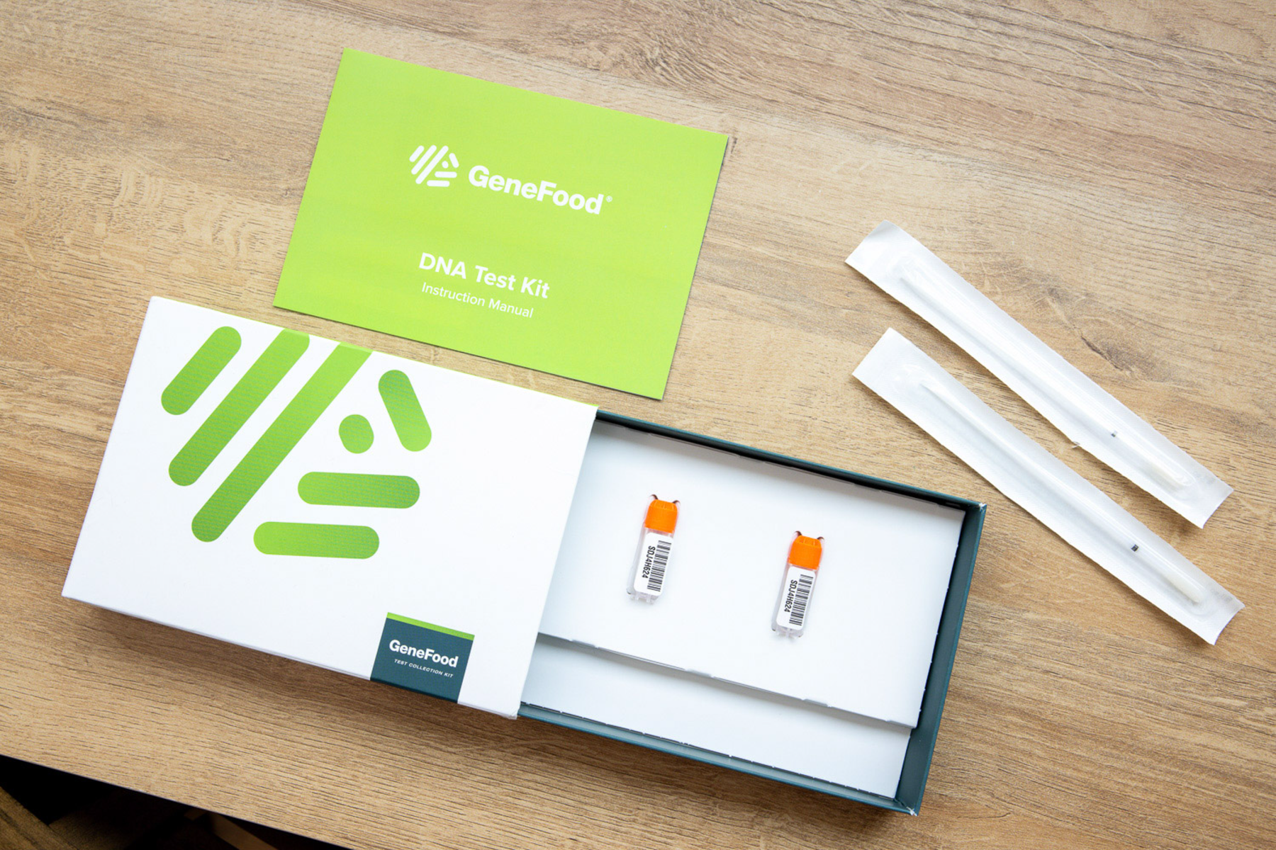 Open GeneFood test kit on a wooden table, showing two labeled sample tubes in the box, the instruction manual, and two sealed cheek swabs